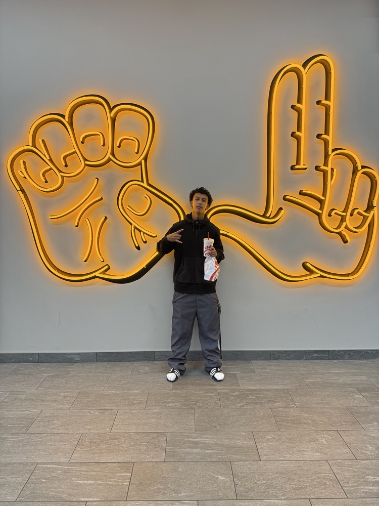 A boy in a black shirt poses in front of a large neon yellow hand design on a wall.