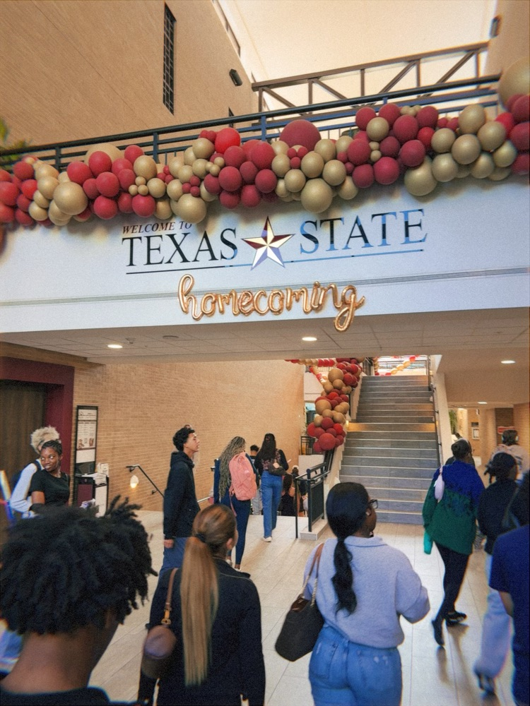 Red, white, and gold balloons hang over entrance. People walk through lobby toward stairs. Banner reads "Homecoming."