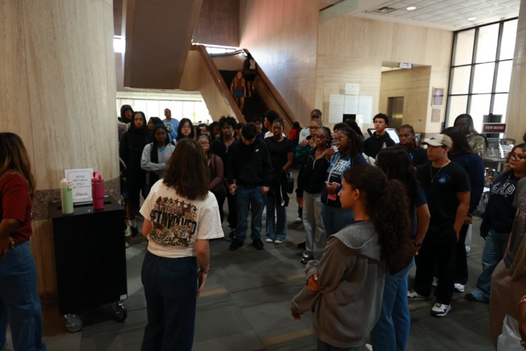 Students stand inside a large building, looking at a table with a water dispenser and cups.