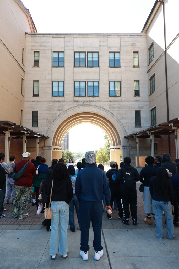 Group of people walking outdoors in line; some wearing hats and sneakers; buildings in background.