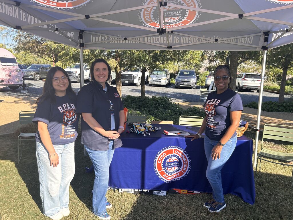 Three women stand under a tent. They wear matching blue shirts and jeans. A table with a blue cover has items on it.