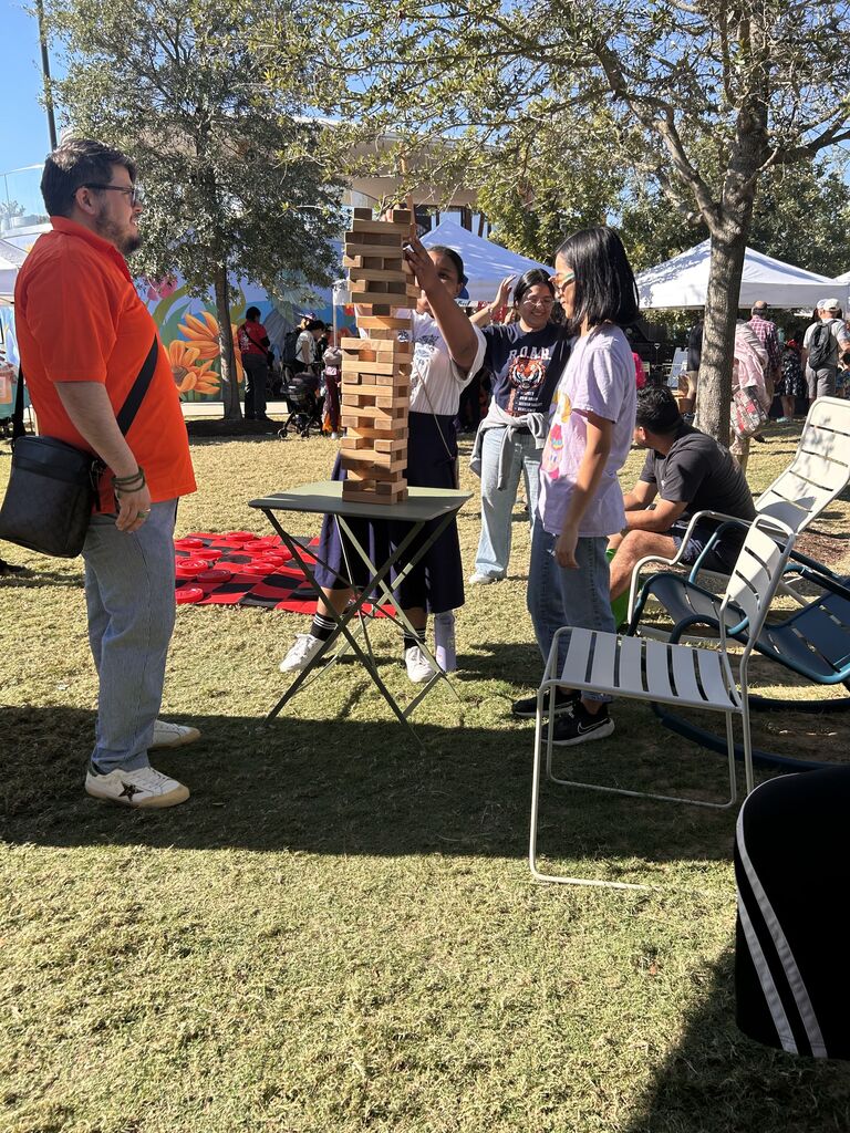 People standing near a tower of wooden blocks on a table. A person holds a block above the tower.