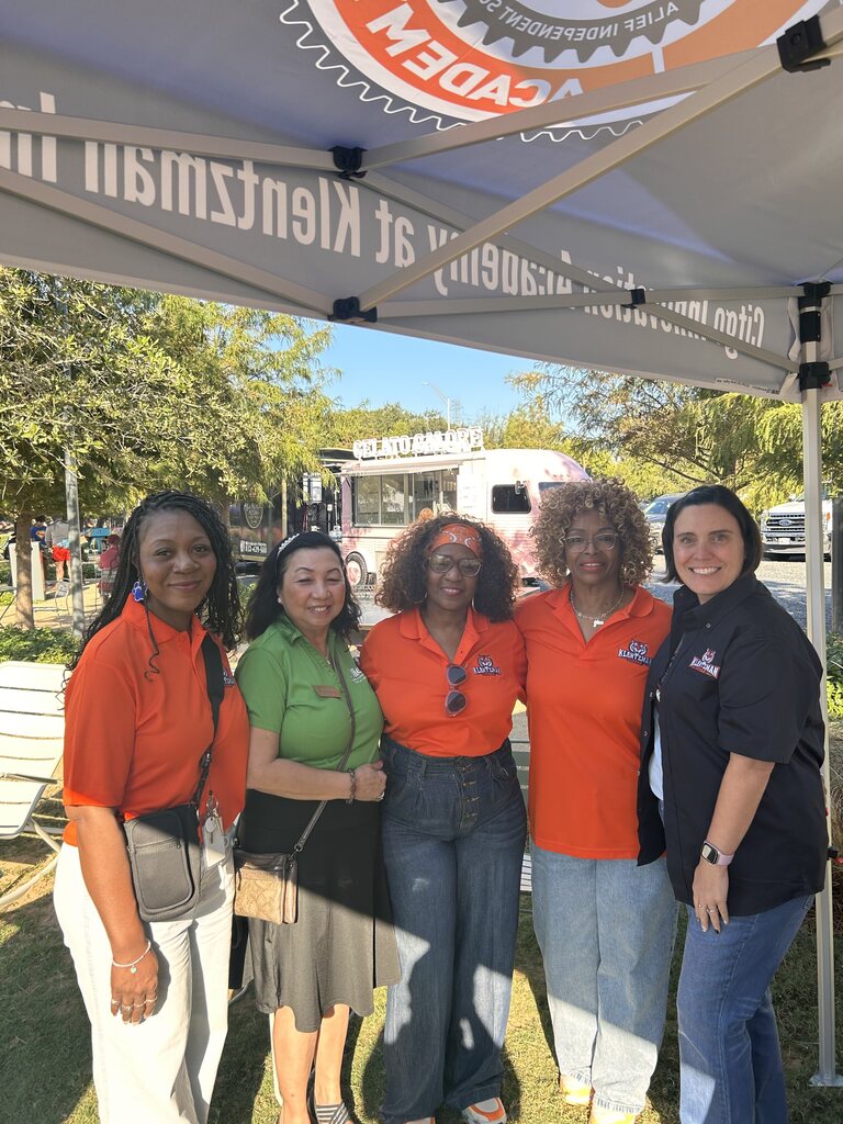 Five adult women in matching orange shirts pose for photo under a tent on a grassy field.