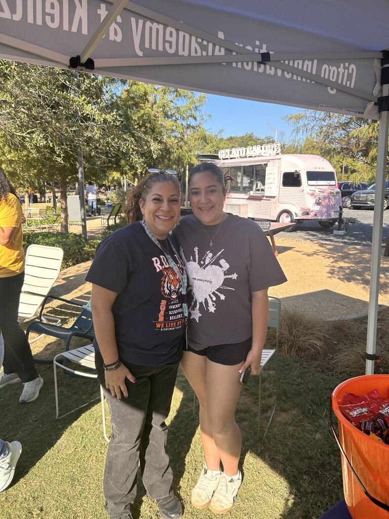 Two women stand under a tent. One wears a gray shirt with a heart design; the other has a necklace. Behind them, a food truck and chairs are visible.