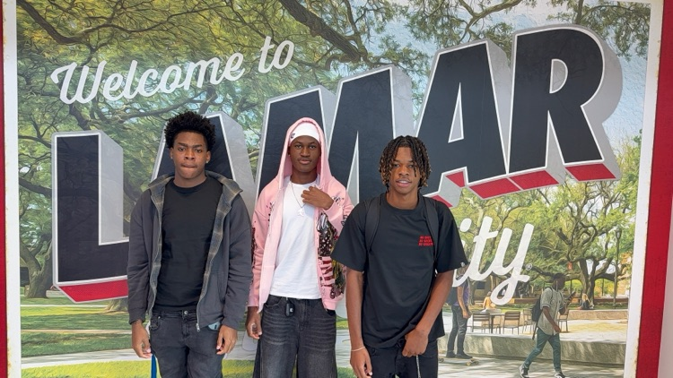 Three students pose for a picture in front of a sign with trees and a park in the background.