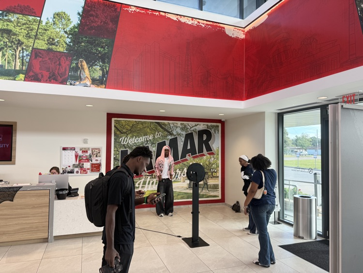 People walking in lobby. Large red wall with large letters. One person carries a backpack. Large framed image on wall.