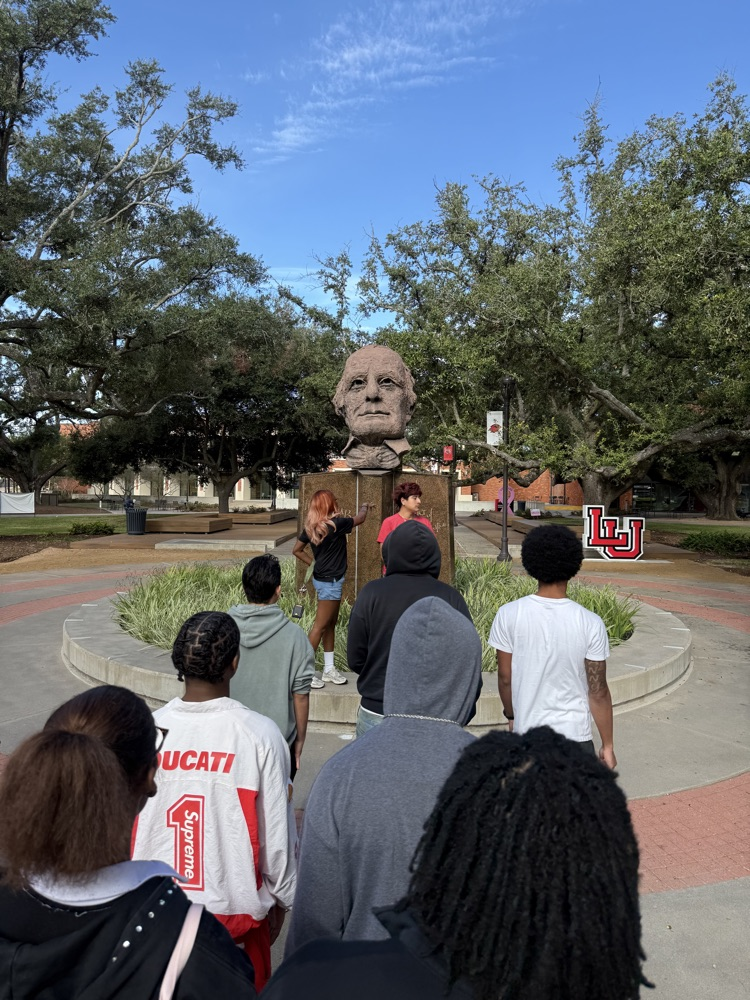 People gather around a statue of a man. A woman stands on top of the statue.