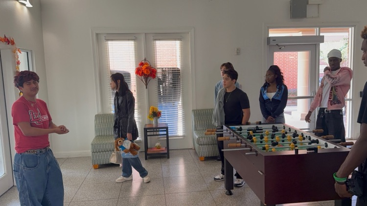 A group of students stand in a room with a foosball table, chairs, and a window.