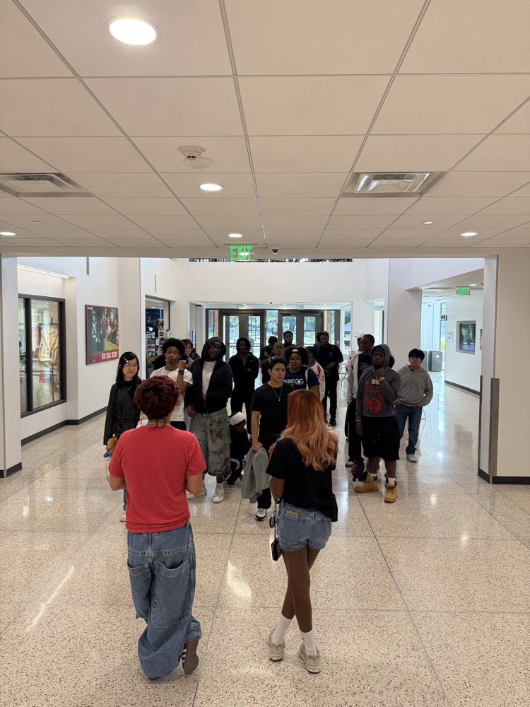 Students walk in hallway with white walls and tiled floor; some carrying bags; framed artwork on walls