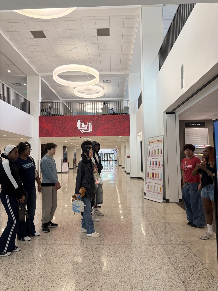 Group of students in a hallway with high ceiling and light fixtures, standing in line and holding bags.