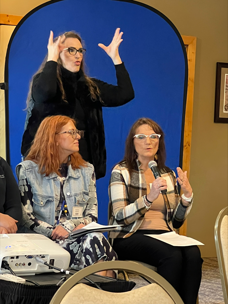 ASL interrupter standing behind red haired woman and Karen, holding a microphone speaking