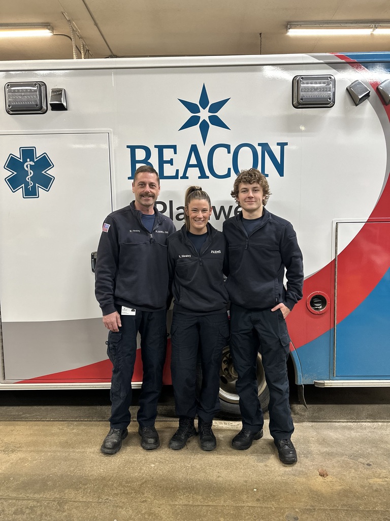 A photo of Nolan Healey and his parents standing in uniform in front of a Beacon Plainwell ambulance.
