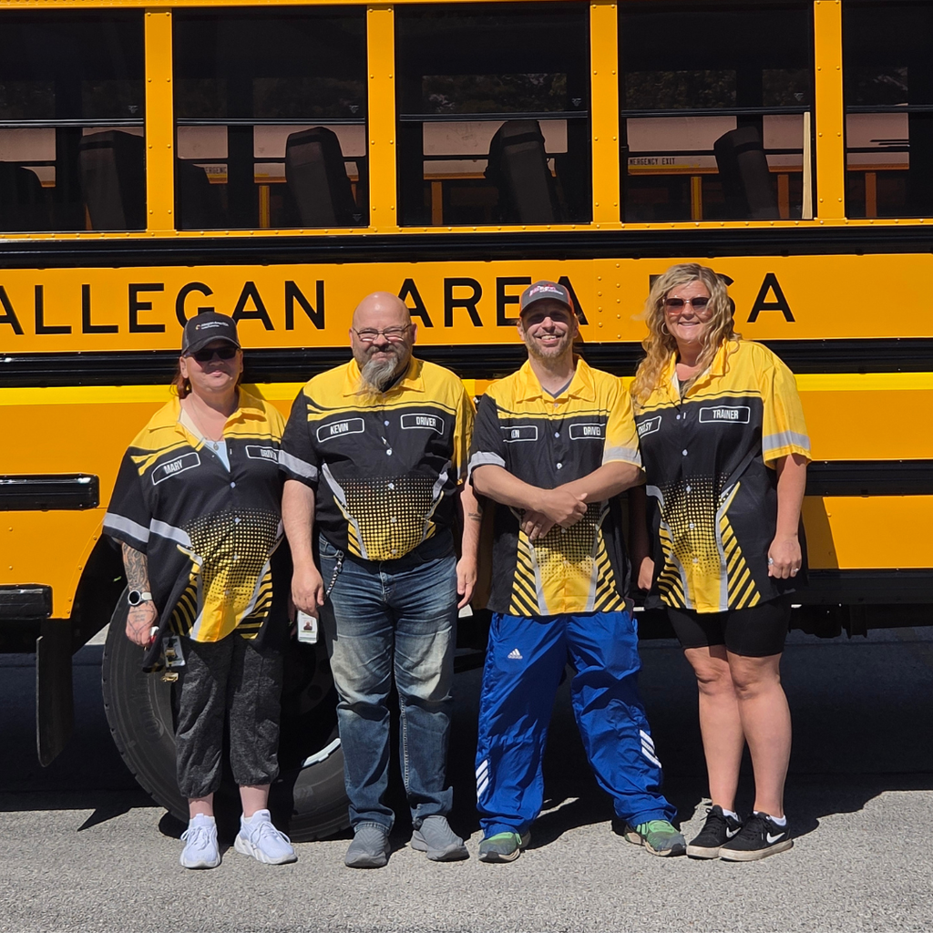 Bus drivers wearing yellow and black matching shirts smile standing in front of an Allegan Area ESA bus.