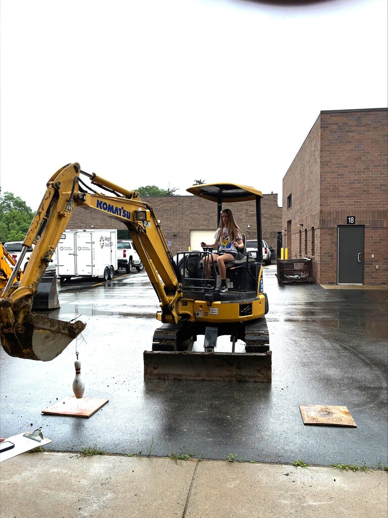Camper on a yellow Komutsu excavator doing challenge testing moving a bowling pin from one target to another.