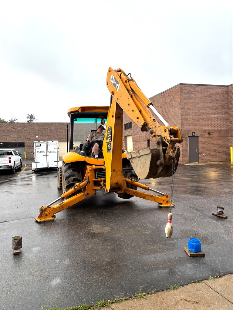Camper on a yellow JCP excavator doing challenge testing moving a bowling pin into a coffee container.