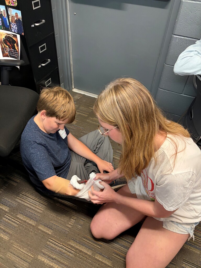 A female camper wraps gauze around a male students arm "injury" that simulates a bone protruding from the skin. 