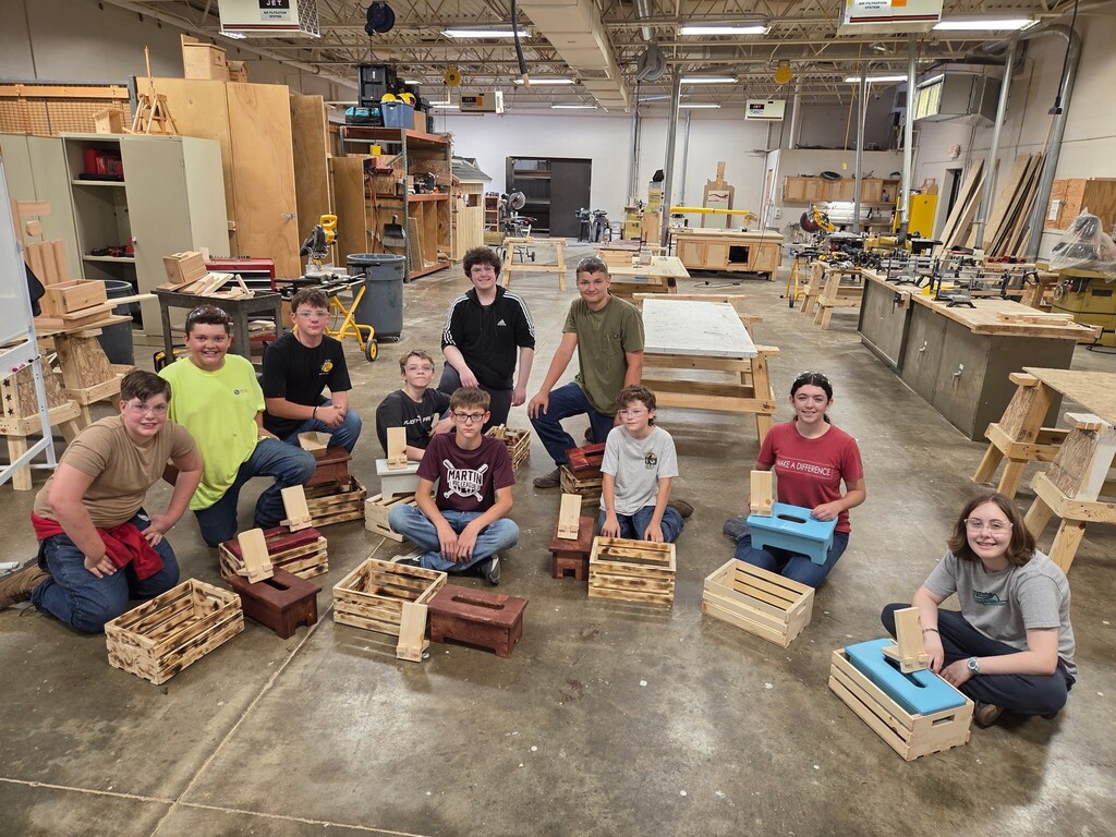 10 male and female campers in the Tech Center construction shop, sitting on the floor in a group for the picture, with all of the items that they made so far.