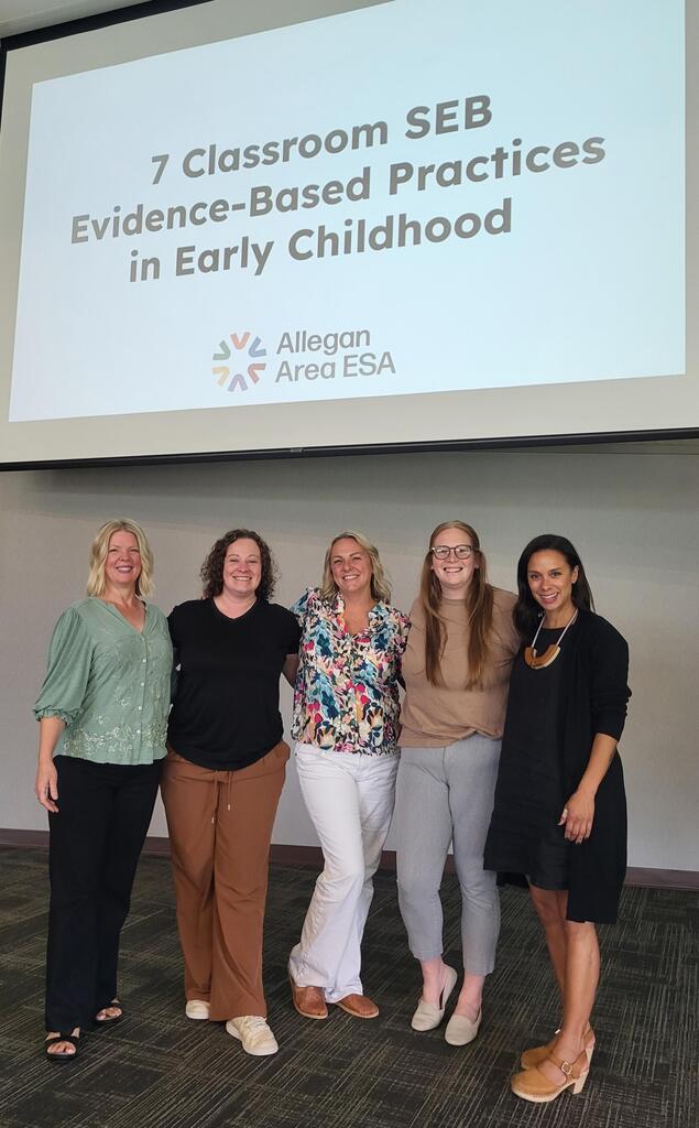 Early Childhood staff members smile in front of a sign that reads "7 classroom SEB Evidence-Based Practices in early childhood"