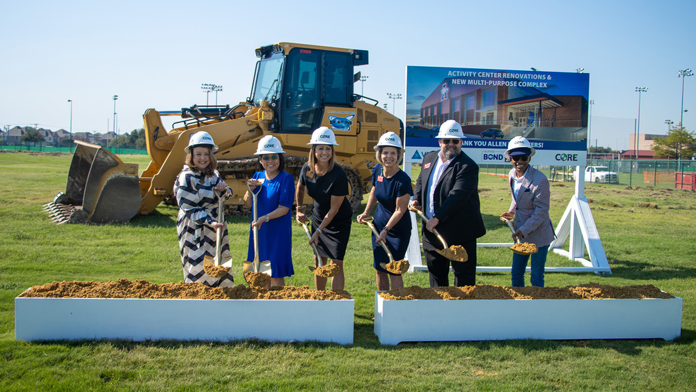 Board of Trustees and Dr. Robin Bullock wearing construction helmets and holding gold shovels pose for a groundbreaking ceremony on a grassy field. A large bulldozer sits behind them, and a sign in the background reads “Activity Center Renovations & New Multi-Purpose Complex. Thank you Allen Voters!” with logos for BCND and CORE. The group is smiling as they turn soil in front of the sign.