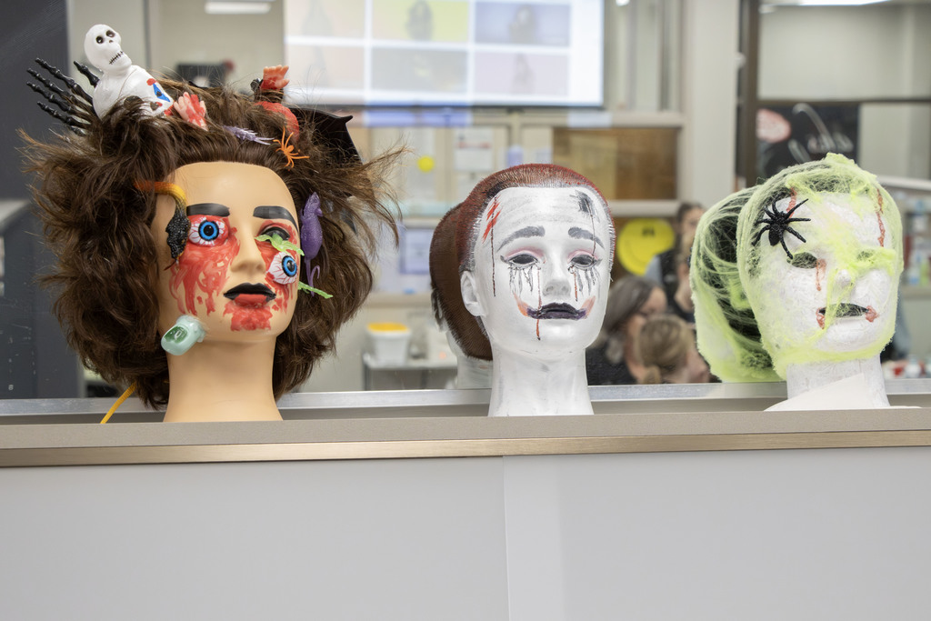 Three mannequin heads decorated with Halloween themes are displayed on a counter. The first head on the left has messy brown hair, fake blood, plastic eyeballs, and small toy insects, spiders, and skeletons tangled in the hair. The middle head is painted white with slicked-back hair, dark dripping makeup around the eyes, and red marks resembling scratches. The third head on the right is wrapped in green webbing with a large fake spider attached and orange streaks resembling blood near the eyes and mouth. The background shows a classroom or salon setting with people working.
