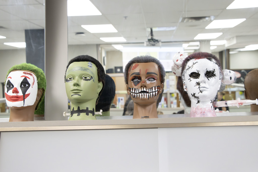 Four mannequin heads with Halloween-inspired makeup are displayed on a counter. From left to right: the first has a white clown face with red lips, black teardrops, and green hair resembling the Joker; the second is painted green with black stitches and bolts on the neck, resembling Frankenstein’s monster; the third has skeletal makeup with black-and-white teeth painted across the mouth and dramatic red eyeshadow; and the fourth is painted white with black cracks and stitches, featuring fake blood and small doll heads attached to the sides, one pierced with a syringe. The background shows a classroom or salon environment.