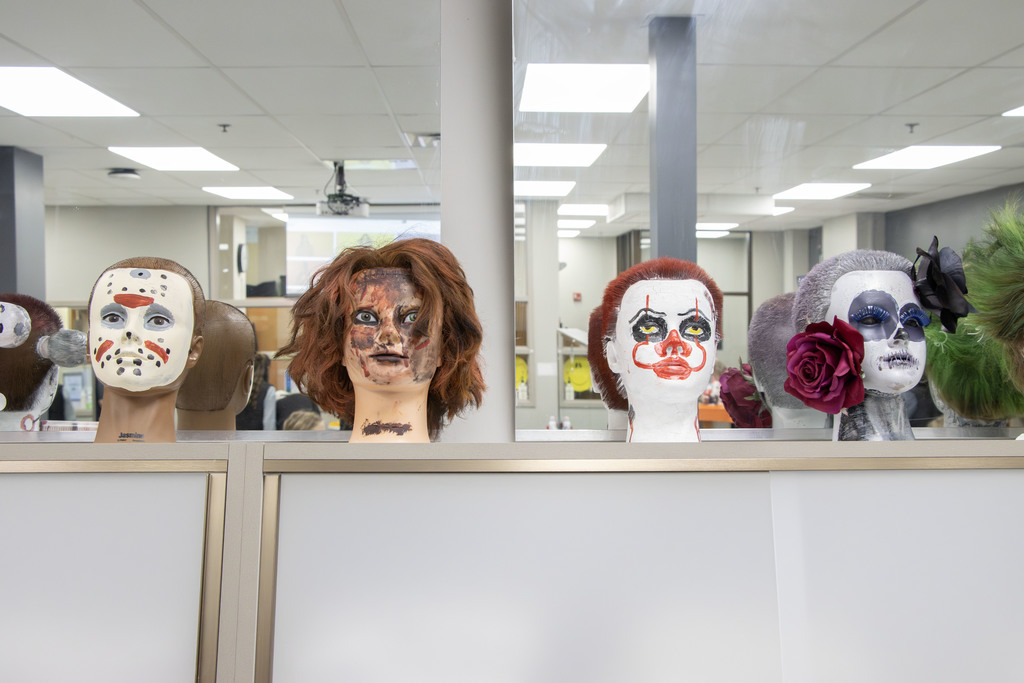 Four mannequin heads decorated with Halloween-inspired makeup and hairstyles are displayed on a counter. From left to right: the first has a white hockey-style mask painted with red and black dots resembling Jason Voorhees; the second has messy reddish-brown hair and a face painted to look scarred and burned, similar to Freddy Krueger; the third is painted white with red clown makeup and yellow eyes resembling Pennywise from It; and the fourth has silver hair styled neatly, a skull design painted on the face, and large red and black flowers attached to one side. The background shows a classroom or salon environment.