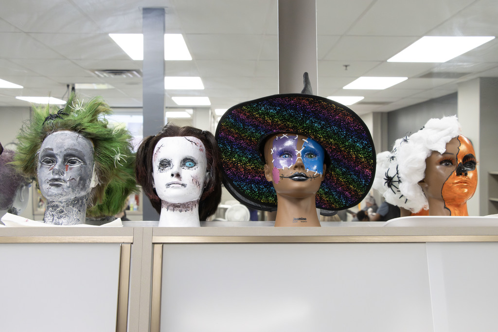 Four mannequin heads decorated with creative Halloween makeup and hairstyles are displayed on a counter. From left to right: the first has messy green and gray hair with fake spiders and a gray-painted face; the second is painted white with dark stitches and pale blue eyes resembling a ghostly doll; the third has bright, colorful makeup with purple glitter around one eye, silver stitches across the face, and wears a large, rainbow-striped witch hat; and the fourth is painted half orange and half white, with cotton-like white hair decorated with fake spiders and a stitched black line down the middle of the face. The scene is set in a classroom or salon space.