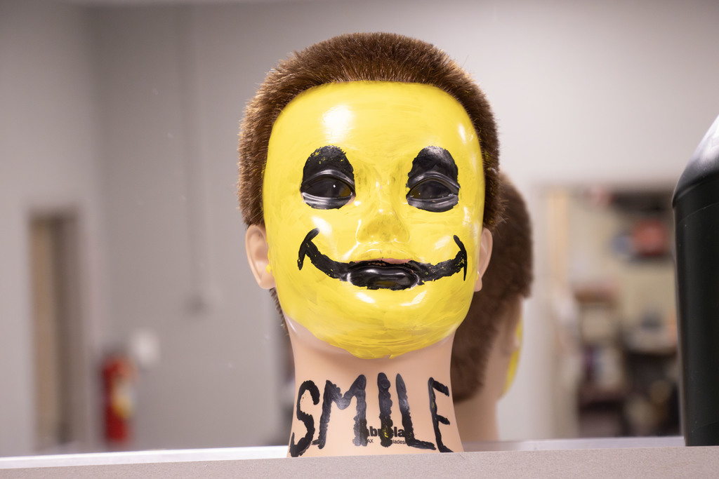 A mannequin head with short brown hair is painted bright yellow with a large black smiley face. The word “SMILE” is painted in black capital letters across the mannequin’s neck. The background shows a blurred classroom or workshop setting.