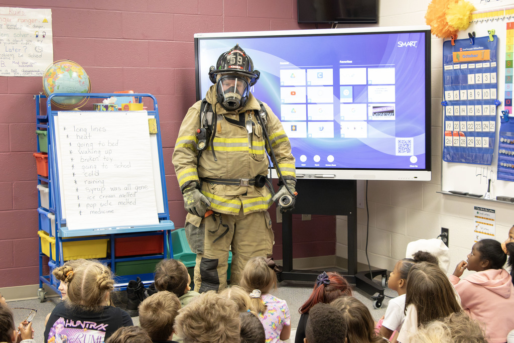 A firefighter wearing full turnout gear, helmet, and breathing apparatus stands in front of a classroom of young students sitting on the floor. The firefighter’s helmet reads “Alliance 59.” Behind them, a SMART Board displays a digital home screen, and to the left, an easel holds a list of words written by students. The classroom is decorated with a calendar, globe, and colorful bins, creating a bright and engaging learning environment.