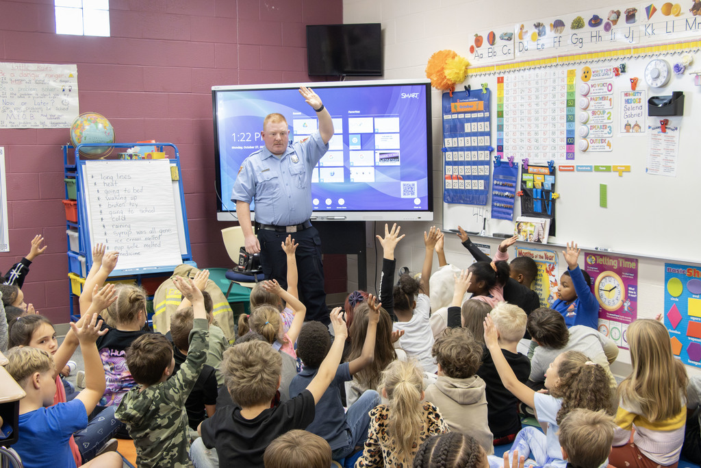 A firefighter in a blue uniform stands at the front of an elementary classroom, raising his hand while speaking to students. The children, seated on the floor, eagerly raise their hands in response. Behind him, a SMART Board displays a digital screen, and an easel with handwritten words sits beside a globe and colorful bins. The classroom walls are decorated with a calendar, alphabet line, and educational posters.