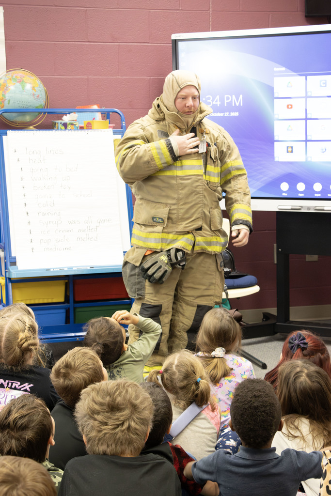 A firefighter in partial turnout gear stands at the front of a classroom, speaking to a group of young students seated on the floor. The firefighter has removed their helmet and breathing mask, revealing the protective hood worn underneath. Behind them, a SMART Board and an easel with a handwritten list of words are visible, along with colorful classroom bins and a globe. The children watch attentively as the firefighter explains the safety equipment.
