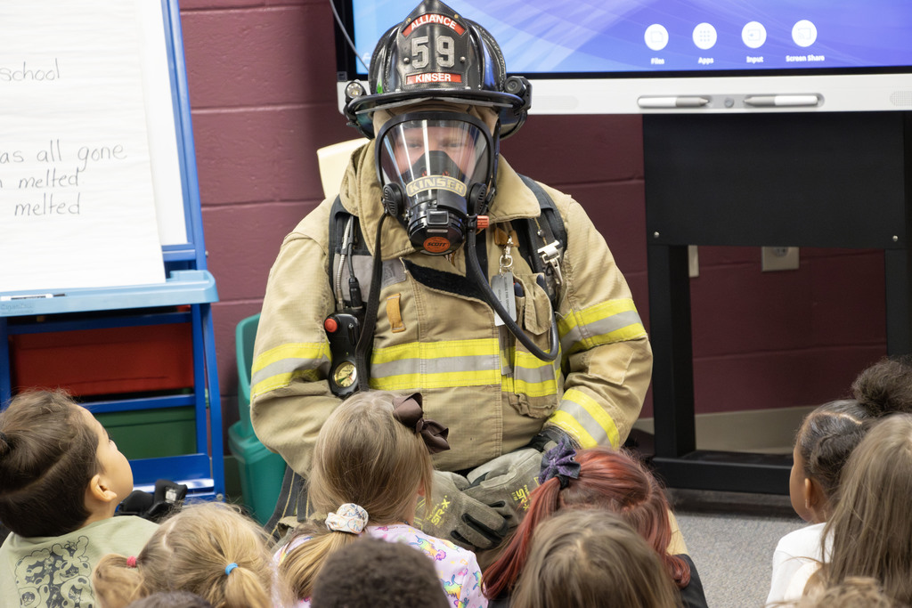 A firefighter wearing full protective gear, including a tan turnout coat, helmet labeled “Alliance 59,” and an air mask, kneels while speaking to a group of young children seated on the floor. The children look up attentively as the firefighter explains safety equipment. The setting appears to be a classroom, with a smartboard, chart paper, and colorful bins in the background.