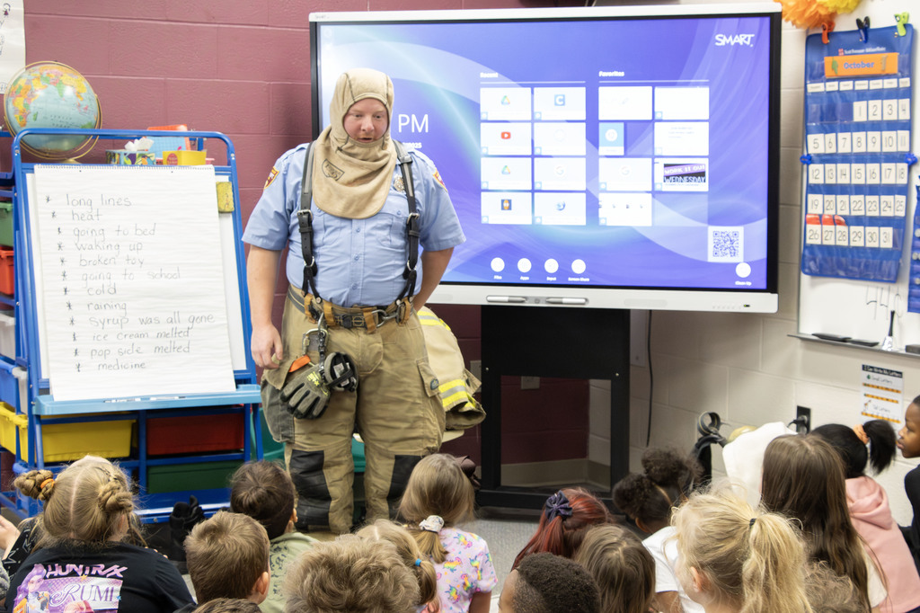 A firefighter stands at the front of a classroom speaking to a group of young students seated on the floor. The firefighter wears a blue uniform shirt, tan turnout pants, suspenders, and a protective hood over their head. Behind them, a SMART Board displays a digital home screen, and an easel beside them holds a list of words written by students. The classroom is colorful and bright, with educational posters, a globe, and a calendar on the wall.