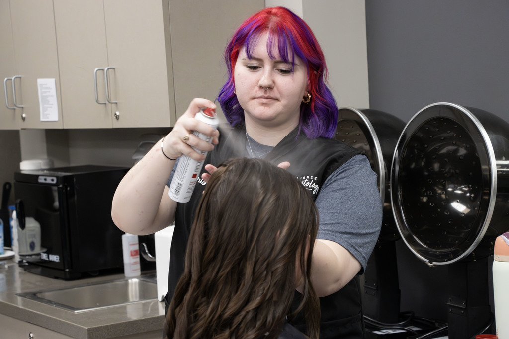 A cosmetology student with bright purple and red hair sprays a client’s styled hair in a salon classroom. The student wears a black vest that reads “Alliance Cosmetology.” Behind them are salon equipment, including two hair dryers, a countertop with supplies, and cabinets. The student appears focused while finishing the client’s hairstyle.