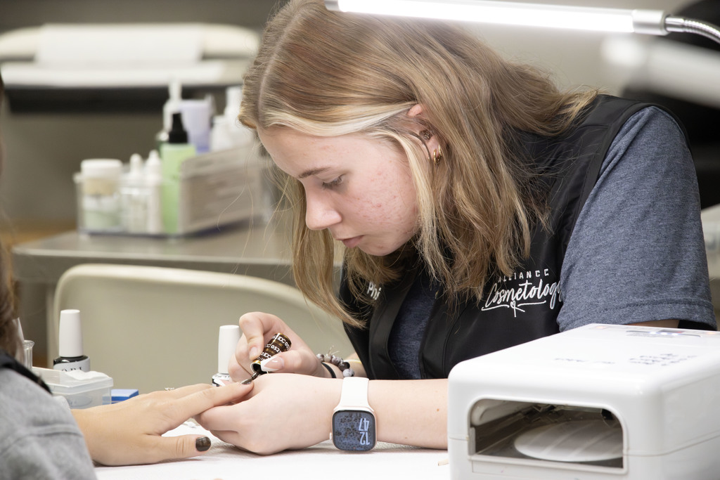 A cosmetology student wearing an “Alliance Cosmetology” vest carefully paints a client’s nails at a workstation. The student focuses intently under a bright desk lamp, surrounded by nail polish bottles and manicure tools. The setting appears to be a classroom salon environment with professional equipment and supplies in the background.
