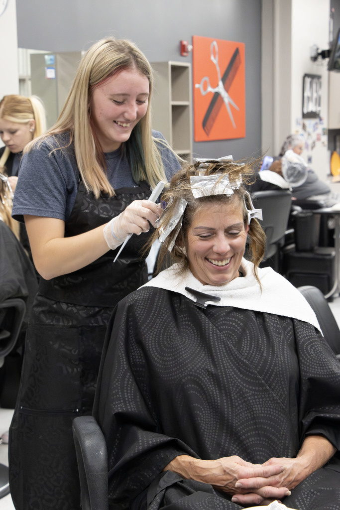 A cosmetology student wearing gloves and an apron smiles while applying hair color foils to a client’s hair in a salon classroom. The client, covered with a black styling cape and a white towel around her neck, is laughing and appears relaxed. In the background, other students work on clients, and a large orange wall art piece featuring white scissors decorates the space.