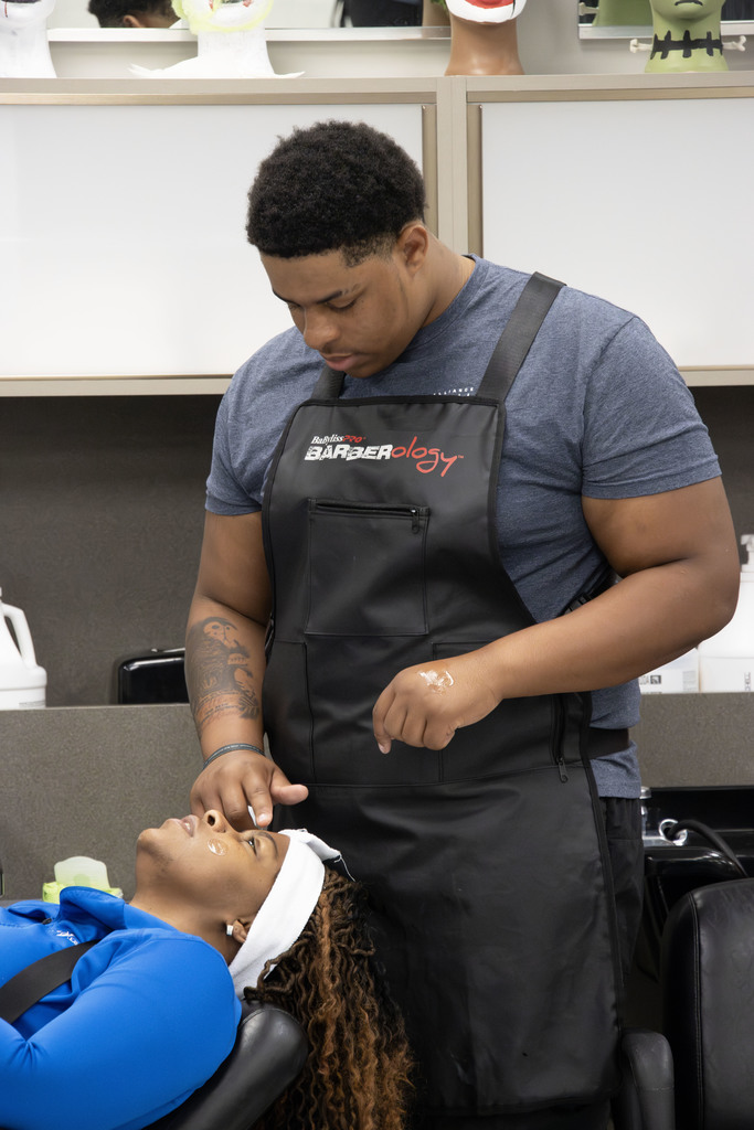 A barbering student wearing a black apron carefully applies facial cream to a client reclining in a salon chair. The client, dressed in a blue top with a white headband, has eyes closed during the skincare treatment. The background shows salon stations with mirrors, equipment, and mannequin heads on display.