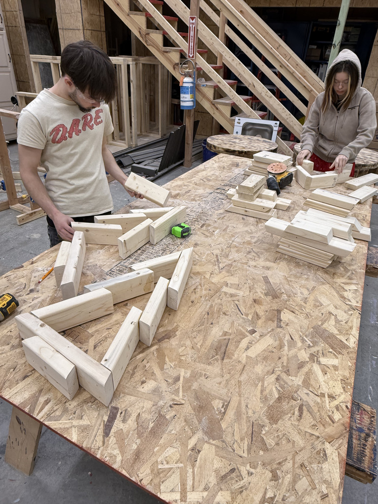 Two high school students work on a woodworking project at a large plywood worktable. The student on the right, wearing a tan hoodie, arranges additional wooden pieces near a power drill and sanding tool. Behind them is a set of wooden stairs, lumber storage, and various tools and materials commonly found in a construction or carpentry workshop.