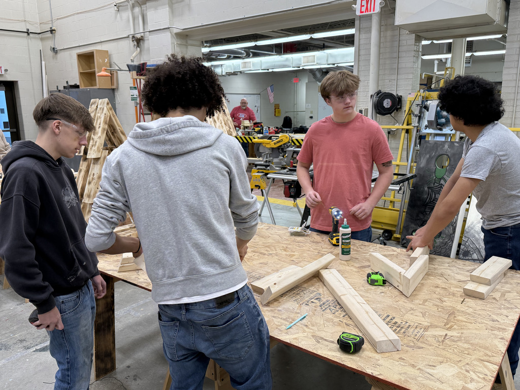 A group of four high school students work together in a carpentry or construction lab. They stand around a large wooden worktable covered with cut lumber pieces, tape measures, a pencil, wood glue, and a power drill. All students wear safety goggles. The workshop around them contains tools, workbenches, and machinery, with additional students and projects visible in the background. The students appear focused as they discuss measurements and assembly steps.