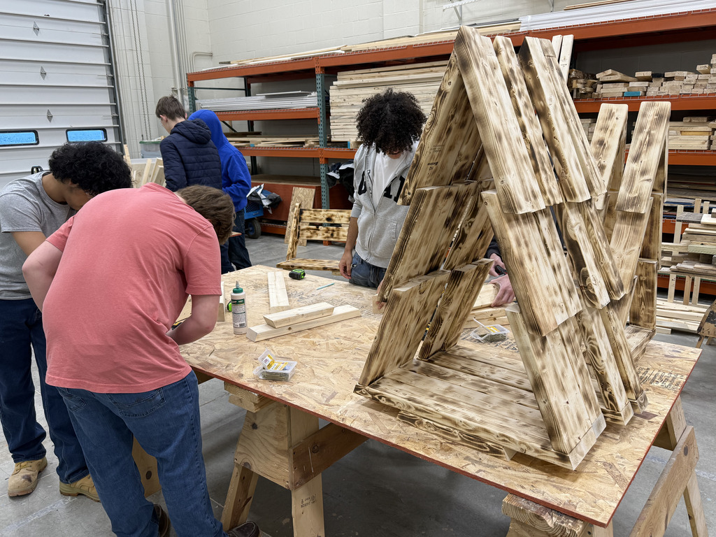 High school students work together in a carpentry workshop, building wooden structures. Several students lean over a large worktable covered with tools, screws, lumber pieces, and a bottle of wood glue. A partially completed A-frame wooden structure made from flame-finished boards sits prominently on the table. In the background, additional students gather around another work area, and shelves stocked with lumber and materials line the workshop walls. The environment is busy and hands-on, with students focused on measuring, assembling, and constructing their projects.