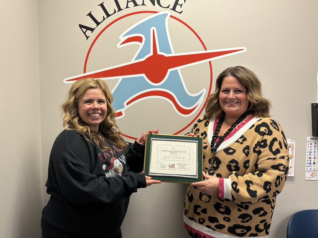 Two women smile while holding a framed certificate in front of an Alliance Aviator logo on a wall. The certificate reads “Hometown Hero of the Month” and recognizes Ashley Peterson. The women stand side by side, each holding one side of the frame.