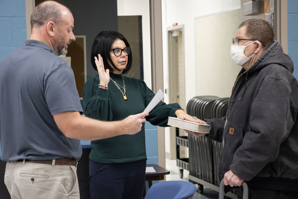 Treasurer Kirk Heath reading the Oath of Office to Teresa Caserta who is raising her right hand with her left hand on the bible while her father holds the bible