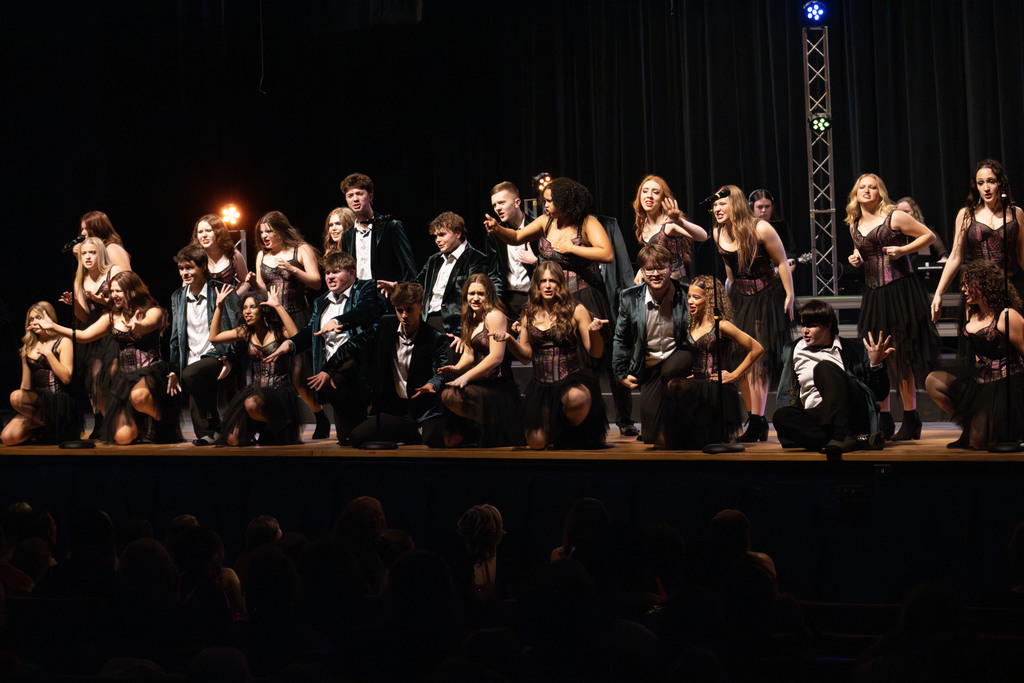 A large high school show choir group performing on stage under bright lights. Students wear coordinated costumes, with some in dark green blazers and others in black skirts with corset-style tops. Many are kneeling at the front with expressive hand gestures while others stand on risers behind them. Stage lighting towers and microphones are visible, and audience silhouettes can be seen in the foreground.