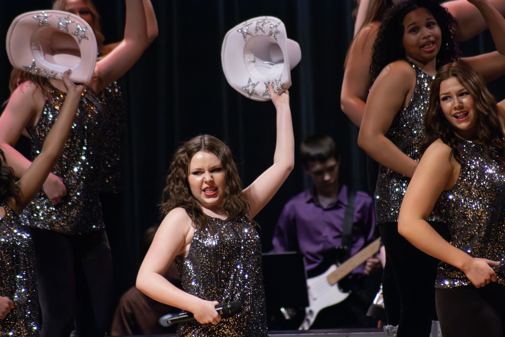 High school show choir students performing on stage in sparkly black sleeveless costumes. A student in the center holds a microphone in one hand and raises a white cowboy hat decorated with silver stars in the other, singing with an expressive face. Other performers around her also hold matching white hats and dance in coordinated poses. A student musician playing an electric guitar is visible in the background against a dark stage curtain.