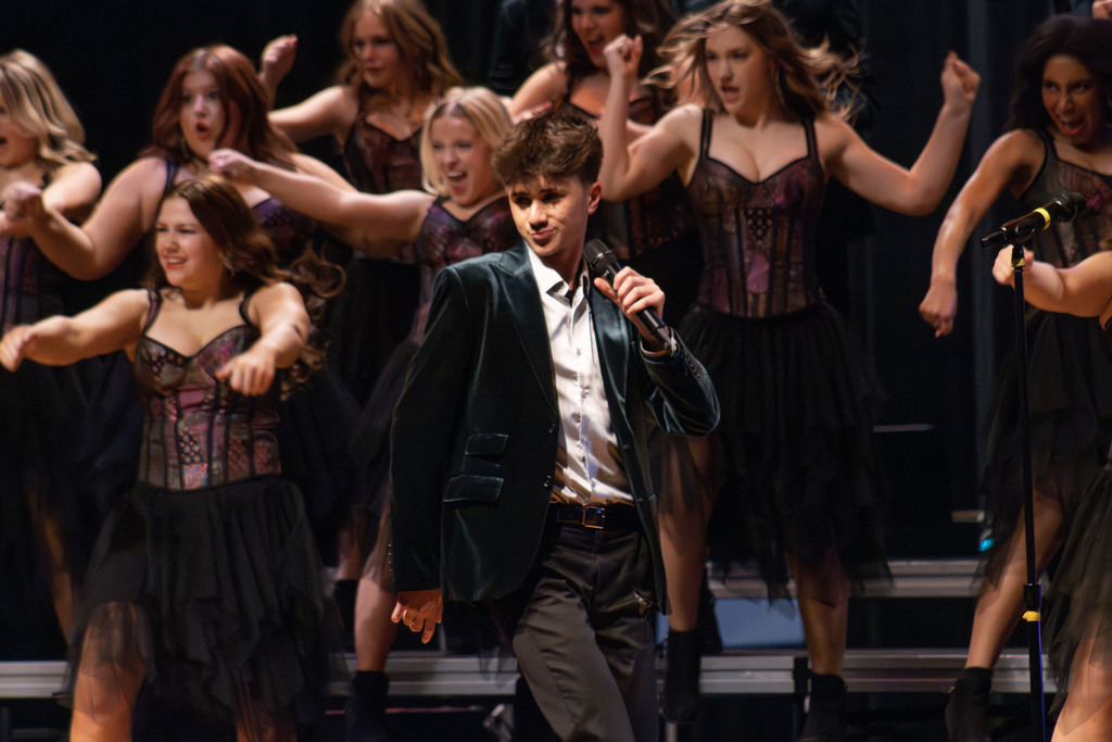 A high school show choir performance on stage featuring a male soloist in a dark green velvet blazer and white shirt holding a microphone. Behind him, dancers in coordinated black skirts and corset-style tops perform energetic choreography with raised arms on tiered risers against a dark stage curtain.