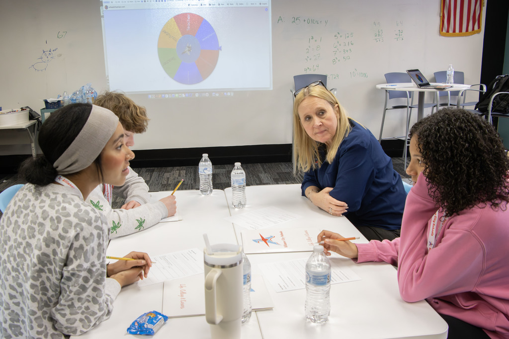 The director of student services sits with three students around a table in a classroom during a small group discussion. Students hold pencils and work on worksheets while listening and talking together.