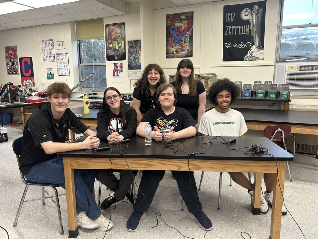 six Alliance High School students and their coach sit and stand around a classroom lab table with quiz buzzers and microphones in front of them, posing for a team photo. The group smiles toward the camera in a science classroom with posters, robotics equipment, and classroom materials visible in the background.
