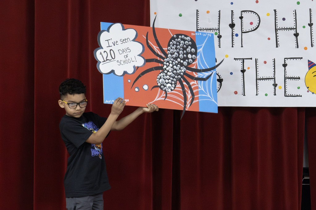 A student standing in front of a sign that says "Hip Hip Hooray It's the 120th day!" holding her project which is a poster board with a drawn on spider that has 120 eyes