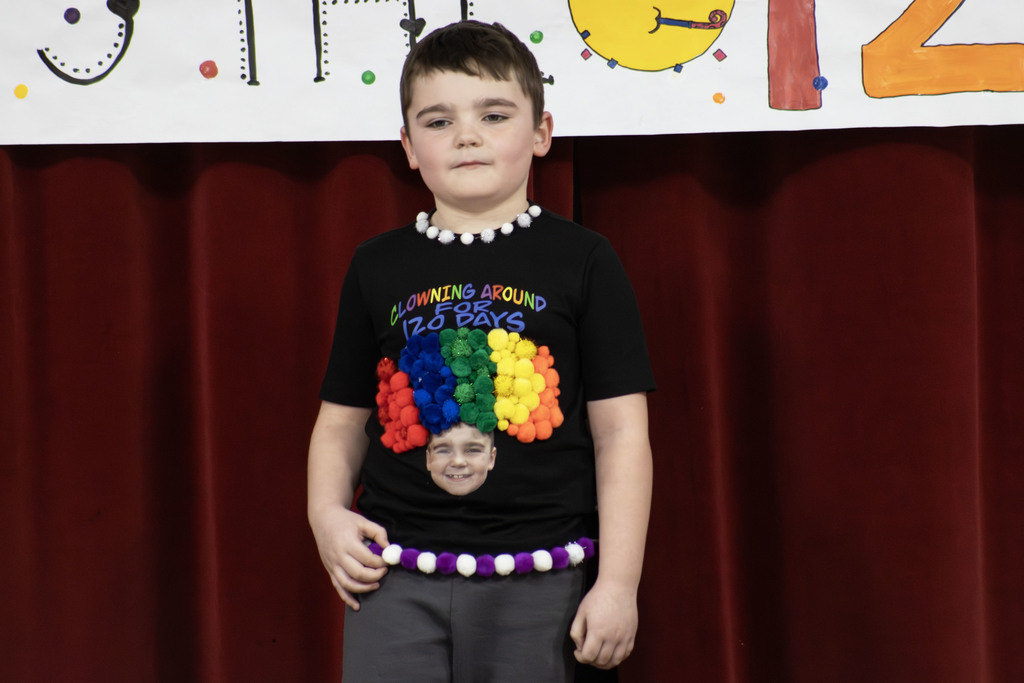 a student wearing a shirt with a picture of himself. He used 120 multi color pom poms that make a clown afro and words that say "Clowning around for 120 days"