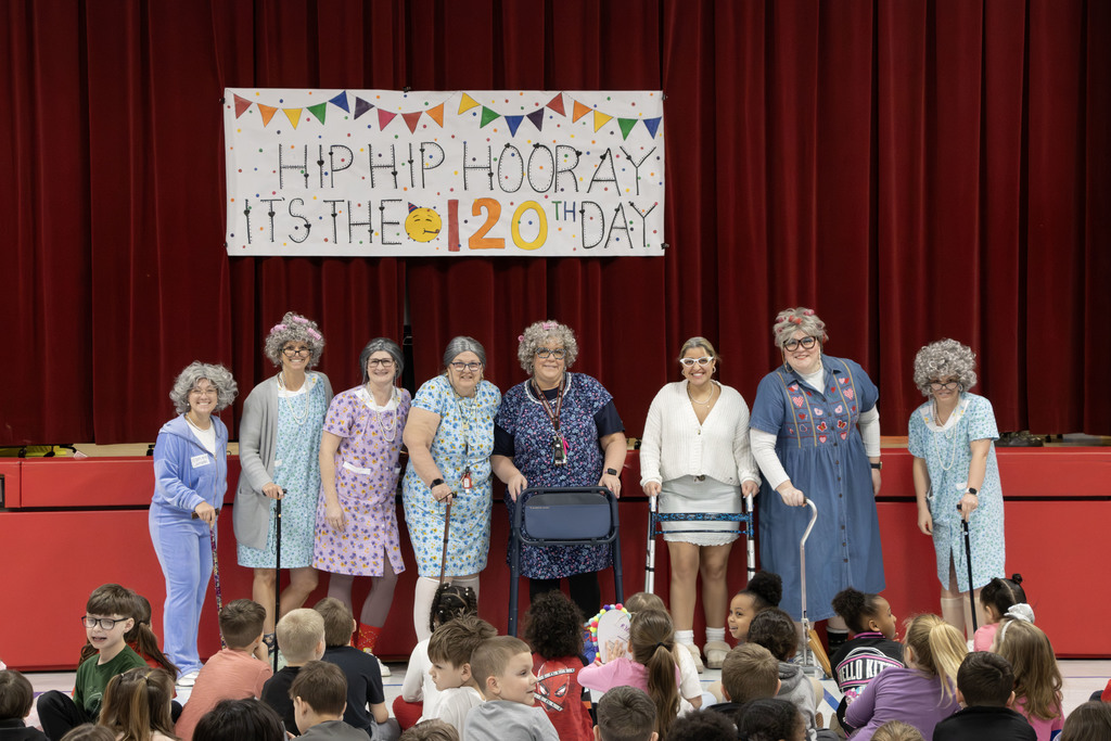 8 first grade teachers dressed as grannies standing in front of the sign that says  "Hip Hip Hooray It's the 120th day!" 